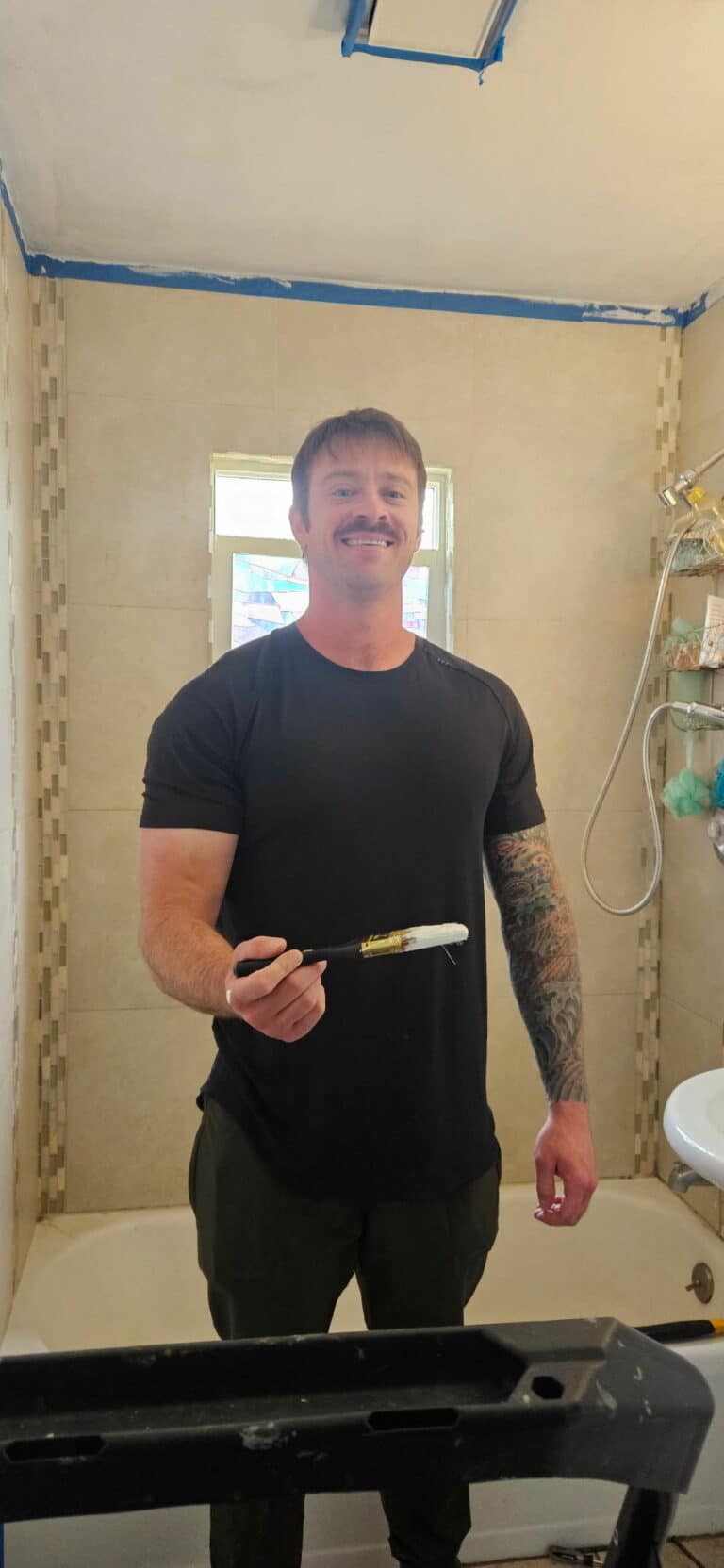 white young adult male with tattoos on his left arm holding a paint brush in a bathroom and smiling at the camera