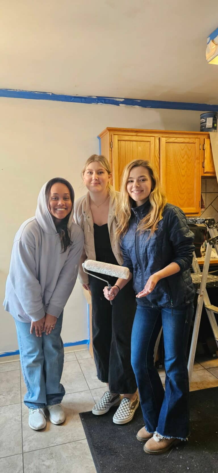 two white young adult females and one black young adult female looking at the camera while standing in a kitchen that they are painting white