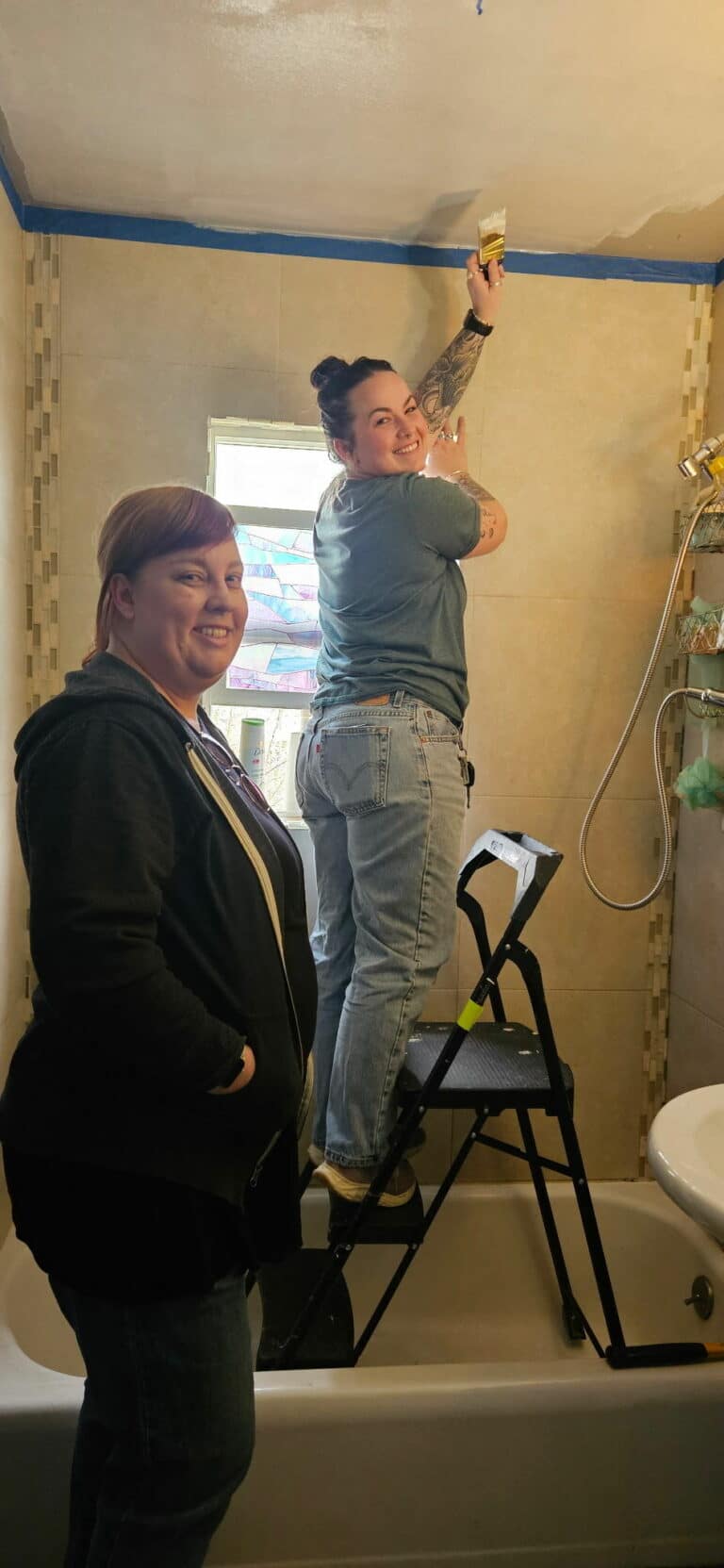 two white young adult females in a bathroom, one standing on a stool, looking at the camera while painting the ceiling with white paint