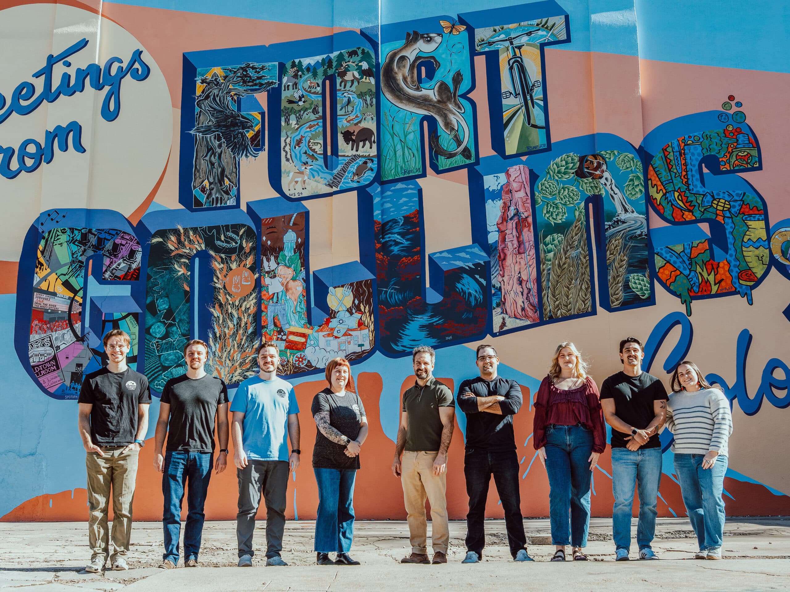 The Beyond Blue Media staff smiling below a mural that says "Greetings From Fort Collins, CO"